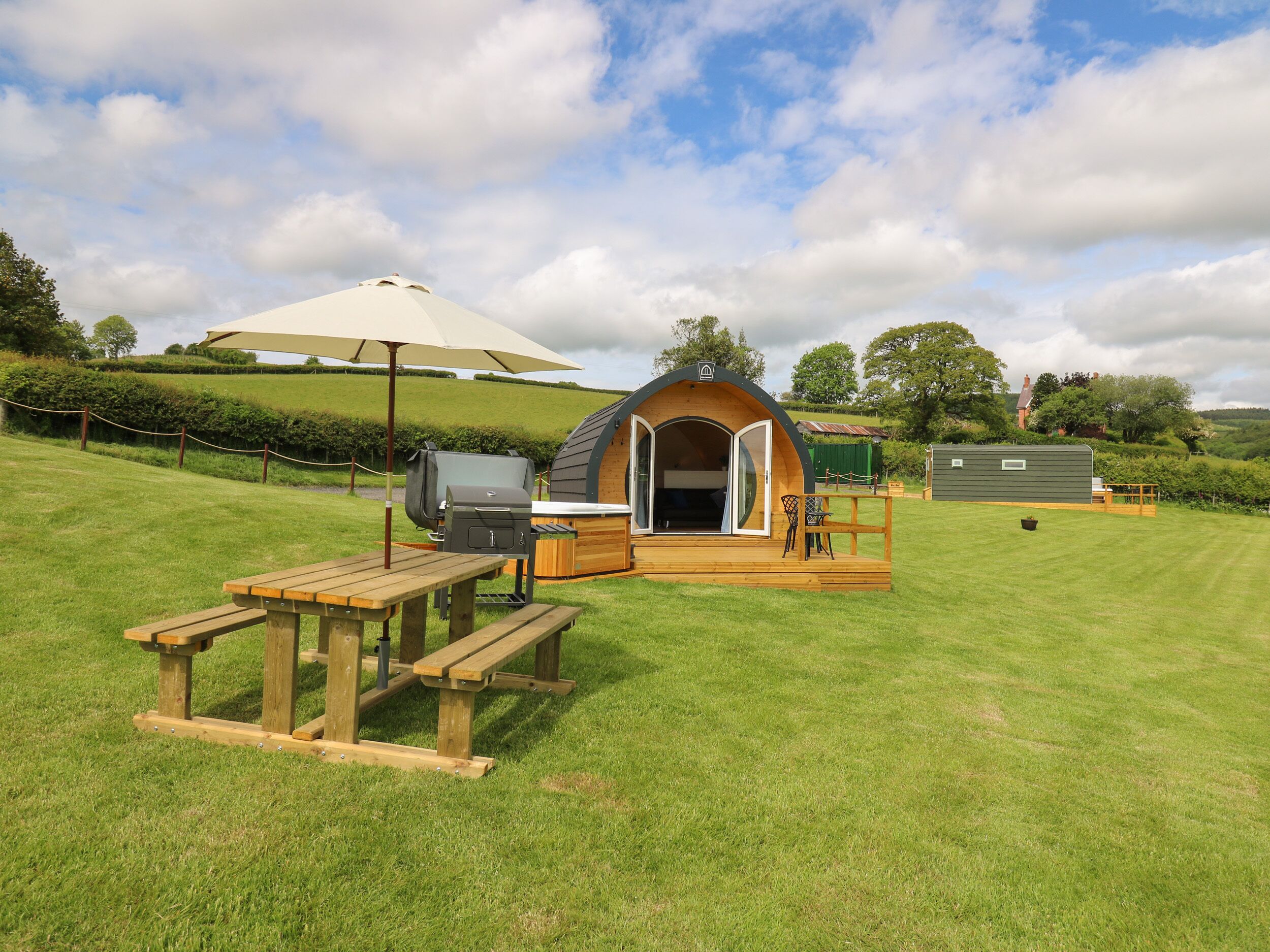 A cozy outdoor accommodation with a wooden deck, picnic table, barbecue grill, and a small covered seating area.