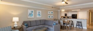 Ein modernes Wohnzimmer mit einem grauen Ecksofa, einem runden Couchtisch und einem Essbereich, im Hintergrund ist eine Küche erkennbar.