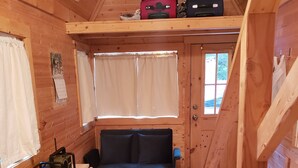 A cozy cabin interior with a wooden floor, a sofa, a small table with a power tool, and a window with curtains. - The Tumbleweed Tiny House (Zirconia)