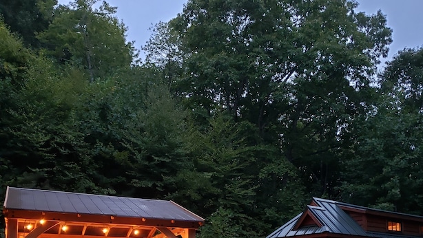 A covered outdoor seating area with chairs and a table, a wooden cabin with a metal roof, and a forested backdrop. - The Tumbleweed Tiny House (Zirconia)
