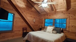 A cozy attic bedroom with a sloped ceiling, a bed with a white comforter, two windows with blinds, and a ceiling fan. - Tranquility at Fall Creek Falls (Spencer)