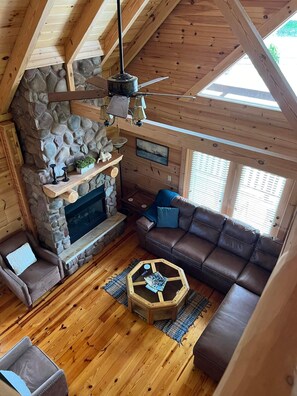 Un interior de cabaña acogedor con chimenea de piedra, vigas de madera en el techo, un sofá de cuero y una mesa de centro hexagonal.