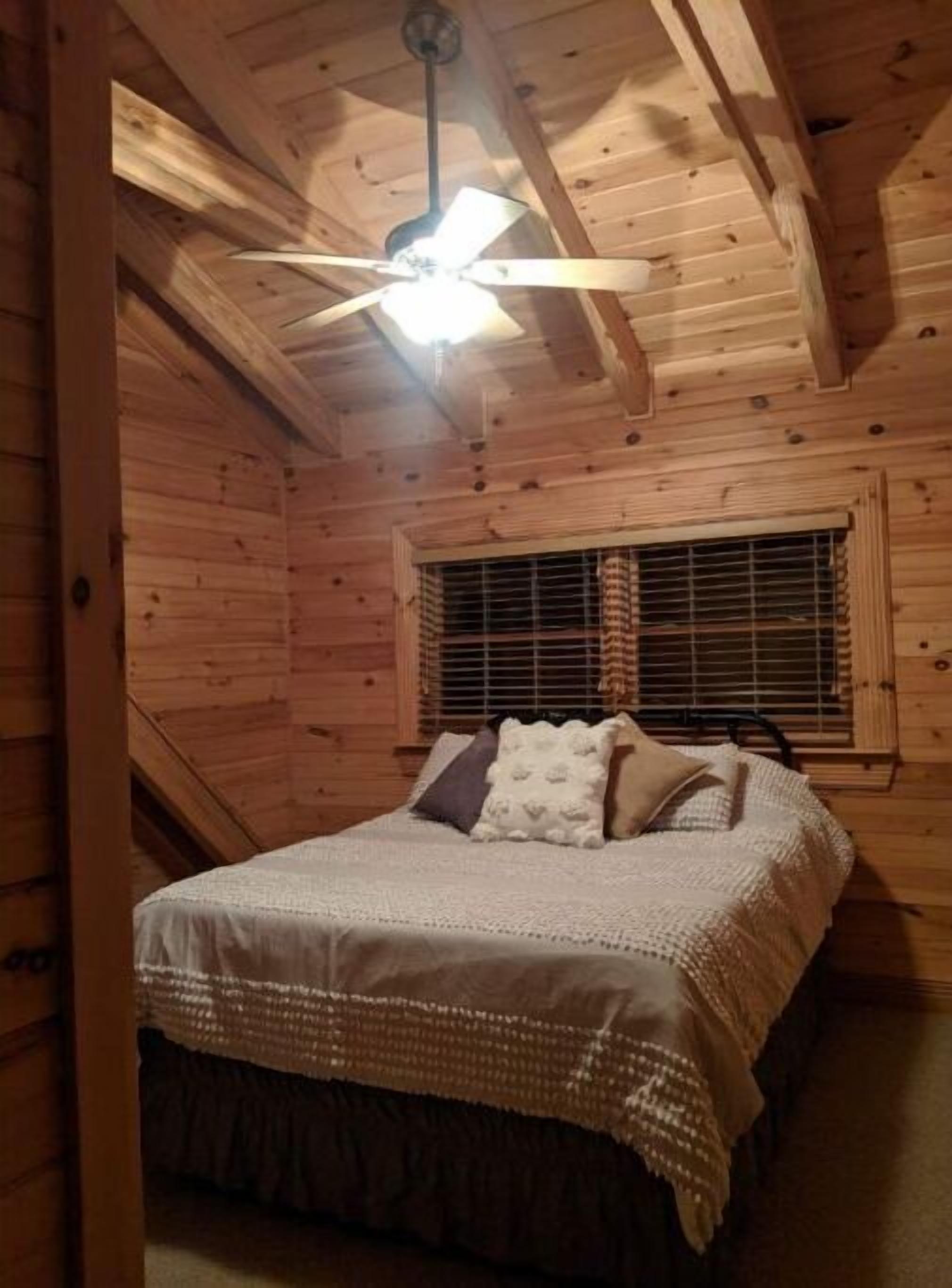 A cozy wooden cabin bedroom with a bed, a window with blinds, and a ceiling fan.