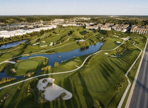 An aerial view of a golf course with water hazards, sand bunkers, and a residential area in the background.