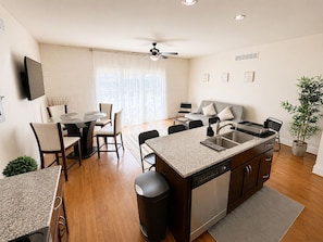 A modern kitchen with a dishwasher, a dining area with a table and chairs, a sofa, and a ceiling fan.