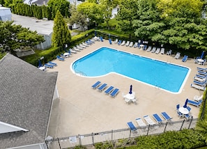A rectangular swimming pool surrounded by lounge chairs and umbrellas, with a fence and greenery in the background.