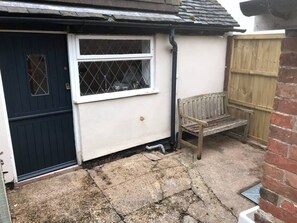 A small house with a dark blue door, a white window with a diamond pattern, and a wooden bench outside. - Village Period Cottage Next to a Idyllic Stream (Coventry)