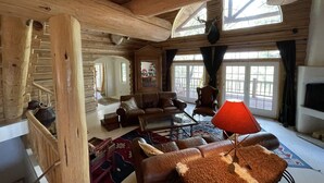 Interior de una cabaña de troncos con una escalera de madera, sofás de cuero, una mesa de centro de vidrio y una lámpara roja.