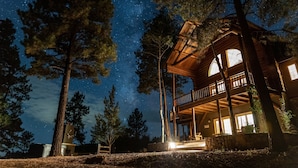 Una cabaña con balcón, rodeada de altos pinos, bajo un cielo estrellado.