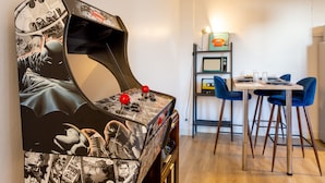 A room with a retro arcade machine, a dining table with chairs, and a microwave on a shelf. - HAPPY KOOL | city center | wifi | private parking (Blois)