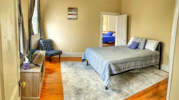 Een slaapkamer met een bed, een stoel, een houten kast en een deur naar een andere kamer.