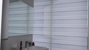 Une salle de bain avec un lavabo blanc, un miroir et une fenêtre équipée de stores horizontaux.