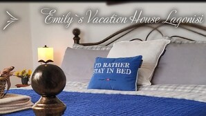Una cama bien hecha con una colcha azul, un almohadón con la frase 'I'd rather stay in bed' y una lámpara sobre una mesita de noche.
