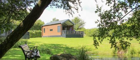 A wooden cabin with a porch, surrounded by a grassy field and a small pond.