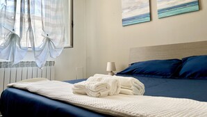 Una cama bien hecha con un cabecero azul, una ventana con cortinas de gasa y dos cuadros de paisajes marinos en la pared.