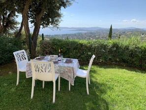 An outdoor dining table with chairs, a bottle of wine, and a bowl of fruit, set against a backdrop of a scenic view of hills and a body of water.