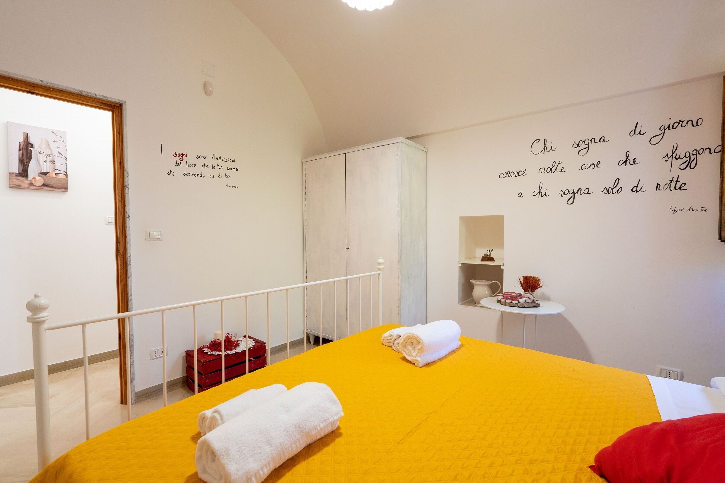 Une chambre d’hôtel avec une couvre-lit jaune, une serviette blanche, un oreiller rouge et un mur orné d’une citation en italien.