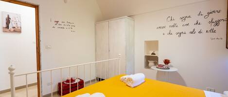 Une chambre d’hôtel avec une couvre-lit jaune, une serviette blanche, un oreiller rouge et un mur orné d’une citation en italien.