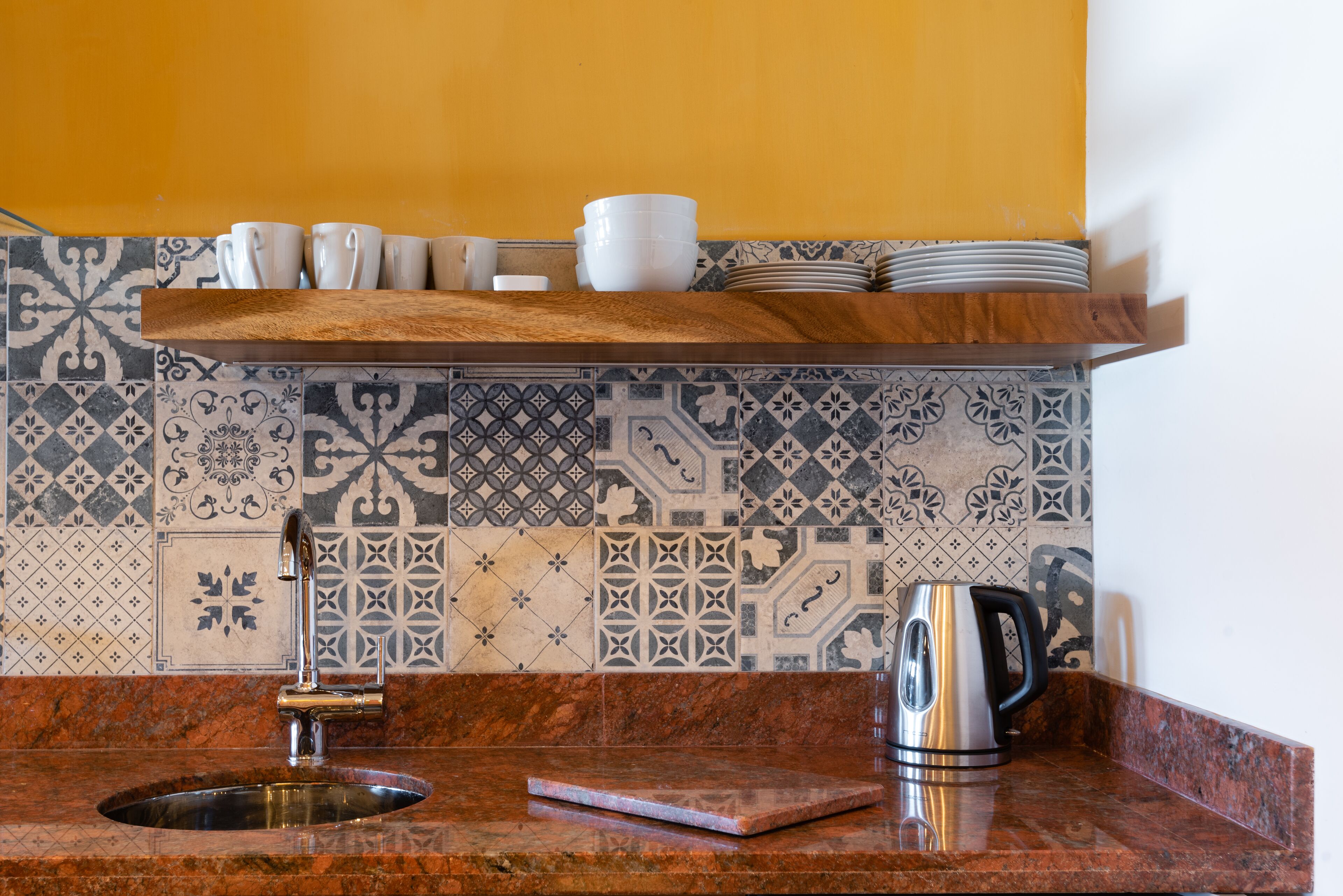 Una cocina con un lavamanos, una cafetera y una estantería con tazas y platos.