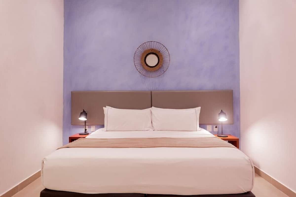A hotel room with a bed, two bedside lamps, a round mirror on the wall, and a headboard.