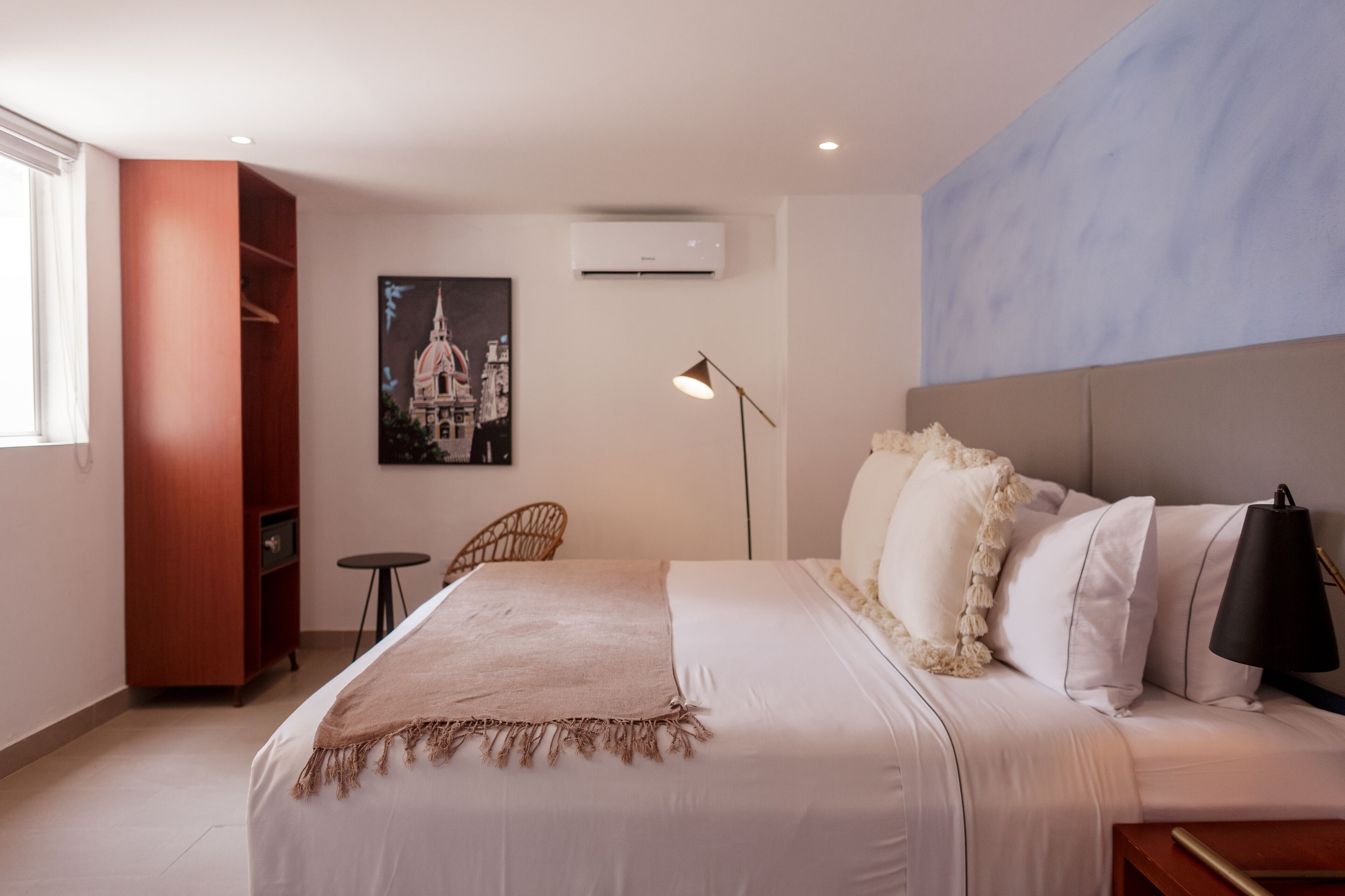 A hotel room with a bed, a chair, a small table, a lamp, an air conditioner, and a painting on the wall.