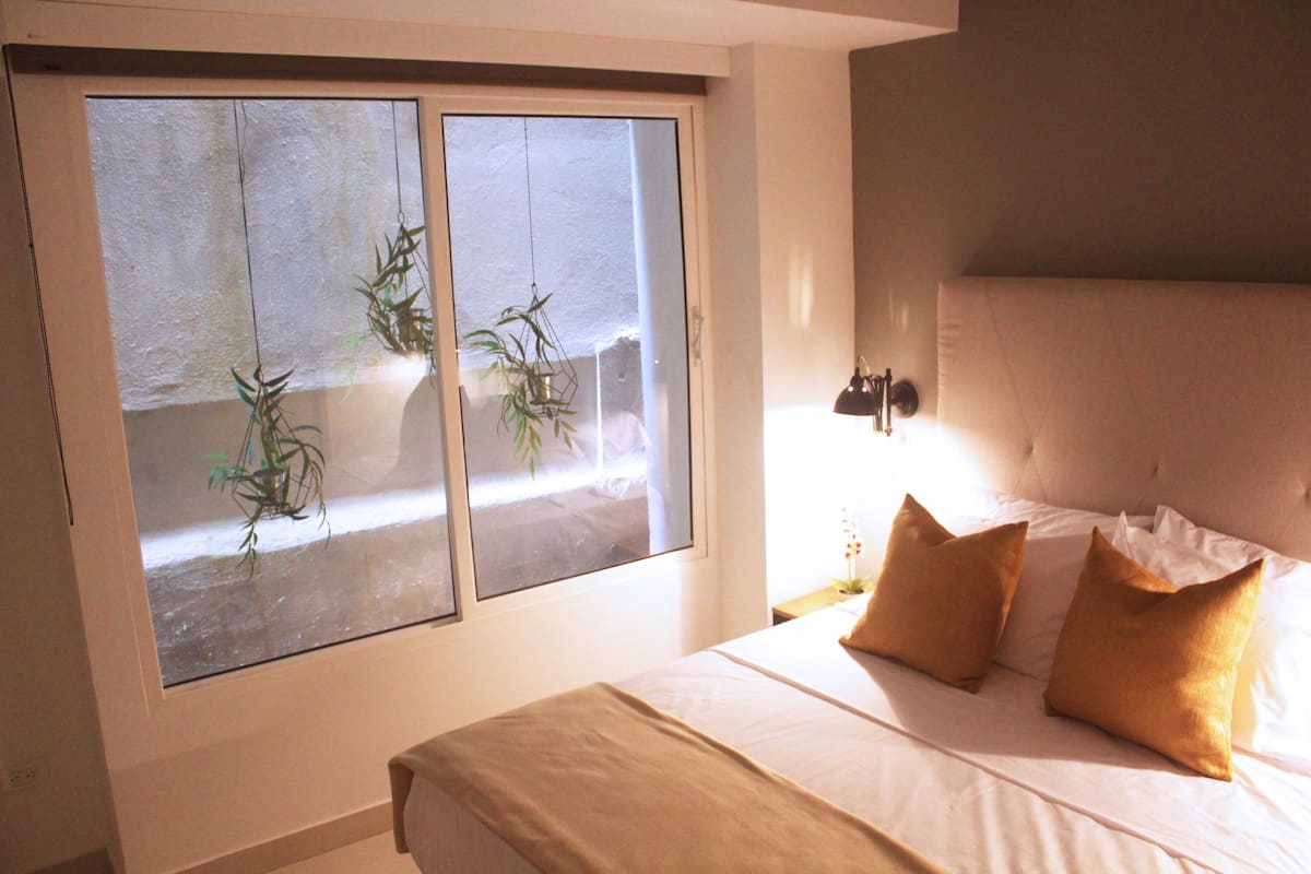 A bedroom with a bed, two pillows, a bedside table with a lamp, and a window with plants.