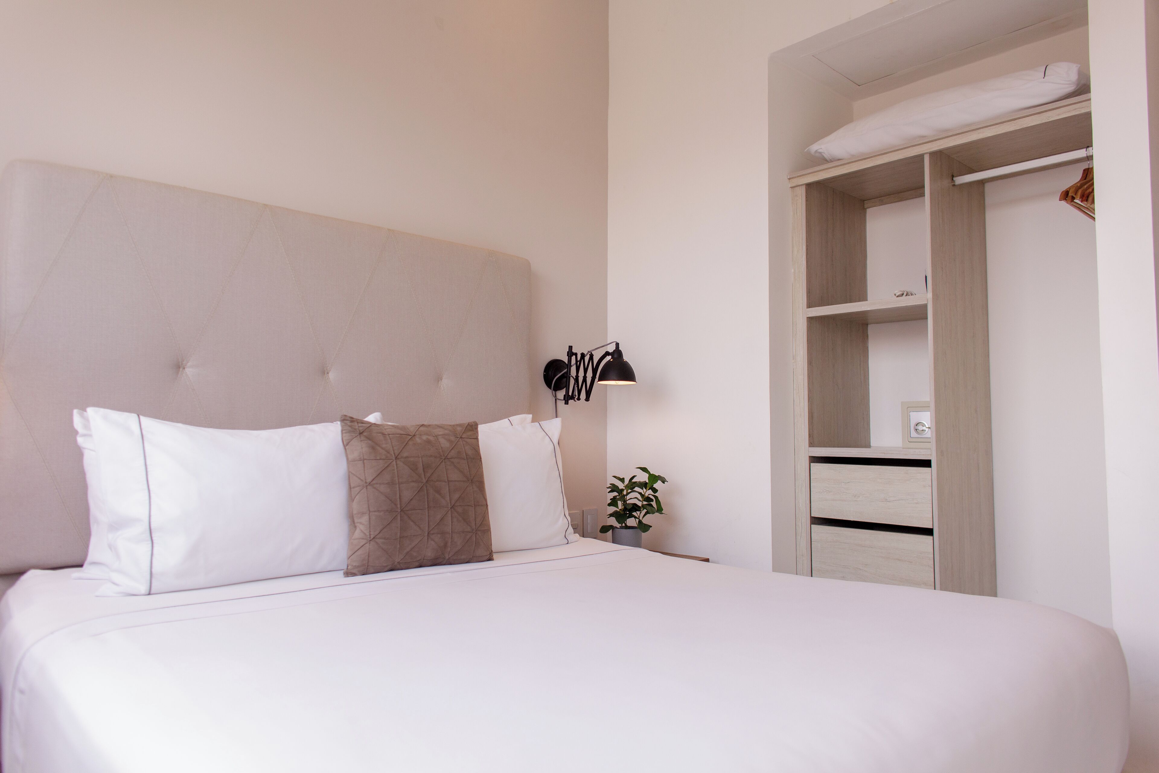 Una cama bien hecha con almohadas blancas y marrones, una lámpara de noche y una pequeña planta en maceta.
