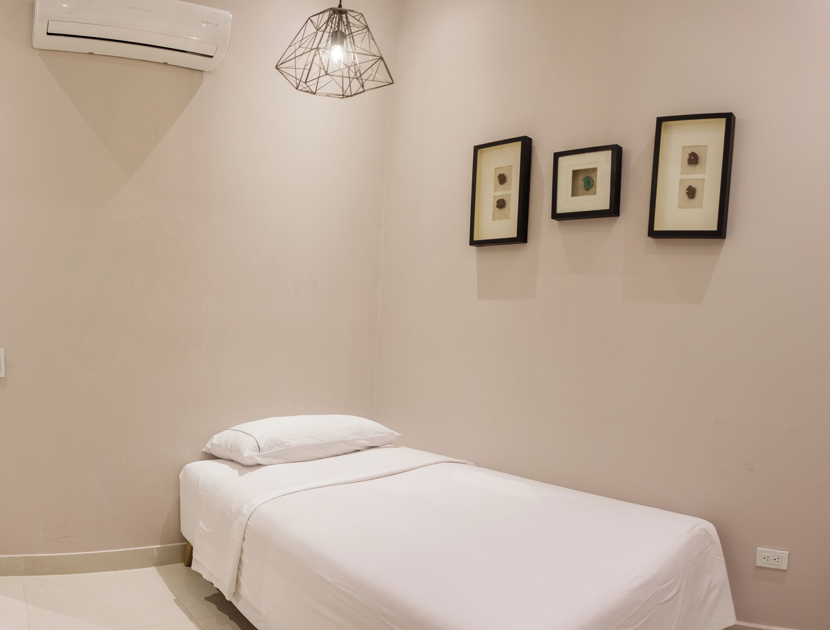 Un dormitorio con una cama individual, un equipo de aire acondicionado, una lámpara colgante de diseño geométrico y tres cuadros en la pared.