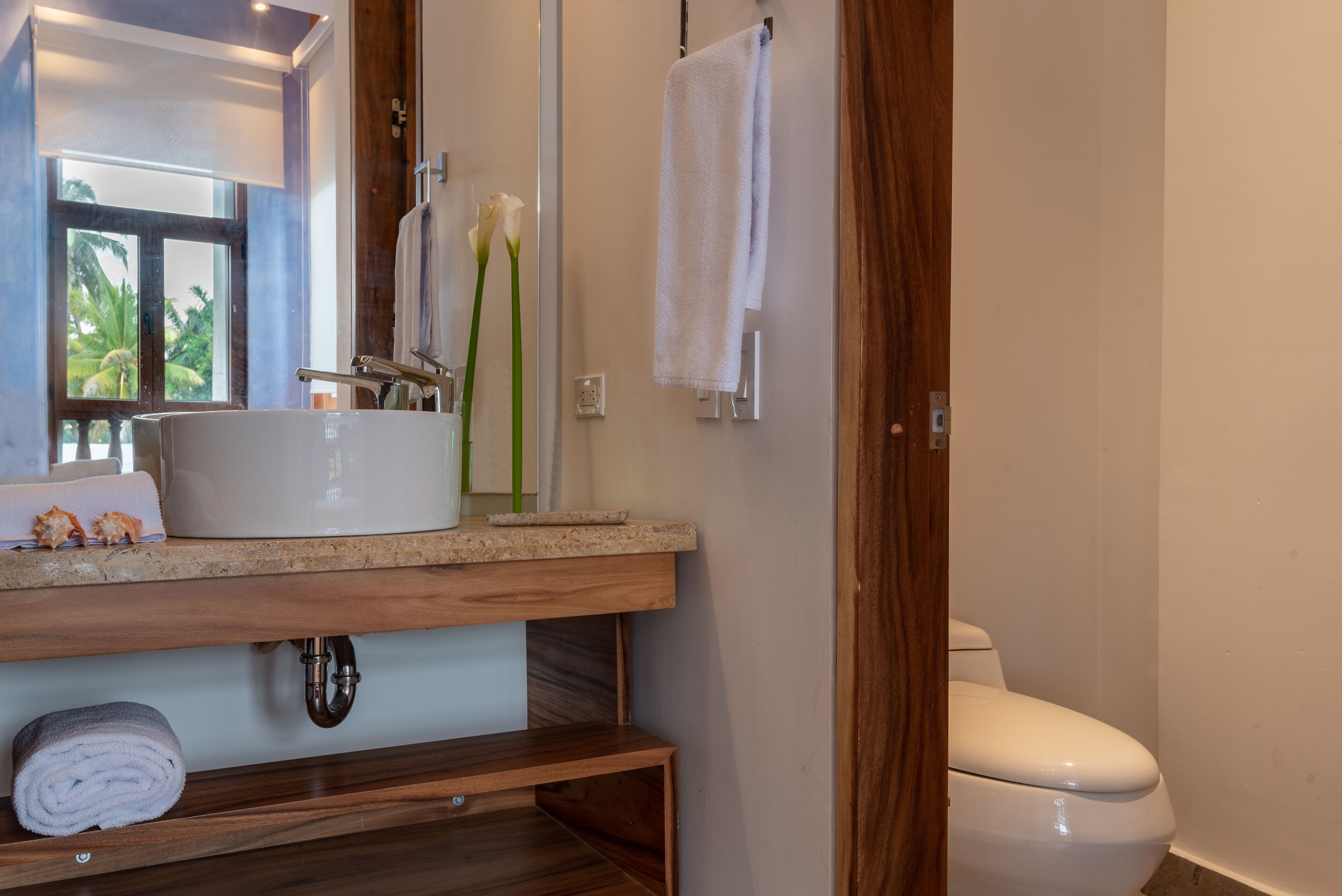 A bathroom with a wooden vanity, a white sink, a toilet, and a window with a view of palm trees.