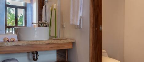 A bathroom with a wooden vanity, a white sink, a toilet, and a window with a view of palm trees.