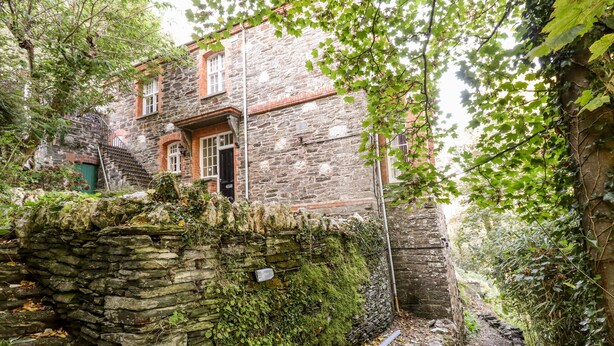 A stone building with a door and windows, surrounded by greenery and a stone wall. - Lewarne (Boscastle)