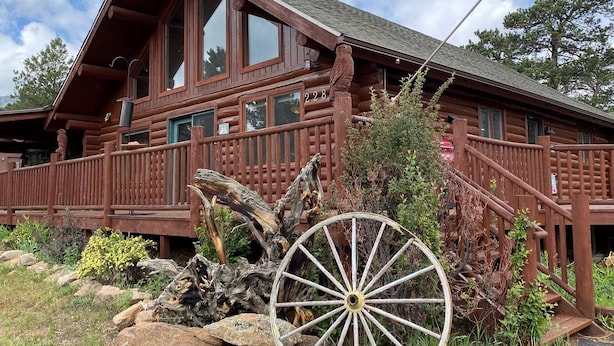 Une cabane rustique en rondins de bois, dotée d’un balcon, de grandes fenêtres et d’une grande roue en bois bien visible au premier plan.