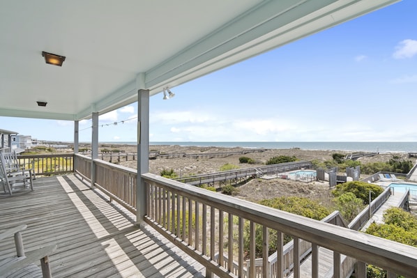 A wooden deck with a railing overlooking a beach and a pool area.