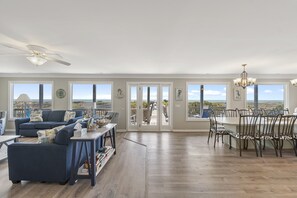 A spacious living room with a large window, a ceiling fan, and a dining area with a chandelier. - "Pharma-Sea West" Oceanfront, Private Pool, 7 BR / 6.5 Bath, PET FRIENDLY (Holden Beach)