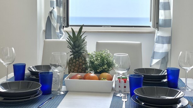 A dining table with plates, glasses, and a fruit bowl, set against a window with a view of the sea.