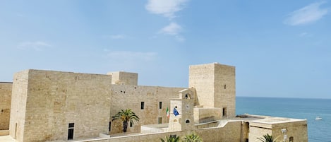 Ein historisches Steinhaus mit einem zentralen Turm, Palmen und Blick auf das Meer.
