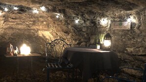 An underground dining area with stone walls, a table with a tablecloth, two chairs, string lights, and lanterns.