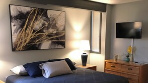 A bedroom with a large bed, a nightstand with a lamp, a dresser, a TV mounted on the wall, and a large abstract painting above the bed.