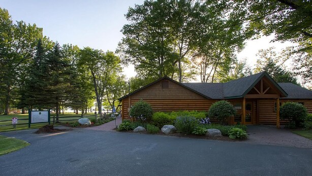 A log cabin with a paved driveway, surrounded by trees and a well-maintained lawn.