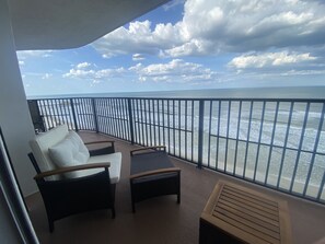 A balcony with two white cushioned chairs and a small table, overlooking a beach with waves and a clear sky.