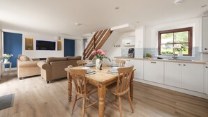 Eine moderne Küche mit Holzausstattung, ein Essbereich mit Holztisch und Stühlen, eine Treppe und ein Wohnzimmer mit Sofa und Fernseher.