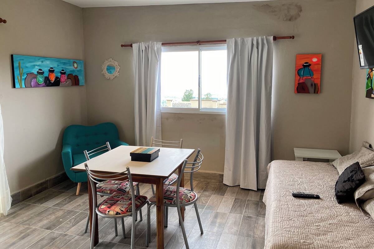 A room with a dining table, chairs, a sofa, a window with curtains, and artwork on the walls.