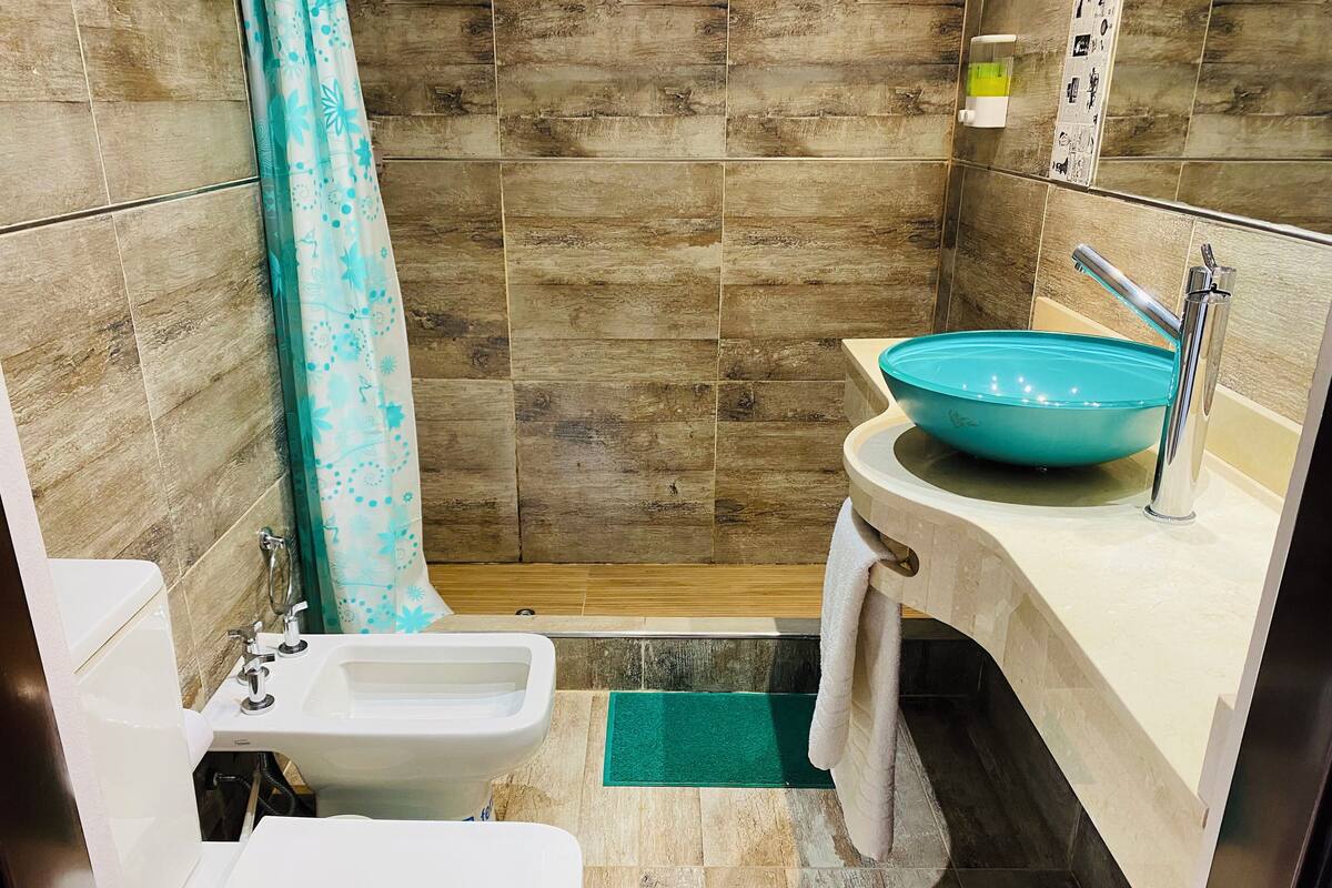 A bathroom with a toilet, bidet, shower, and sink with a blue basin.