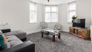 A living room with a grey sofa, a glass coffee table, two chairs, a TV on a wooden cabinet, and large windows. - Luxury 3 bedroom apartment (Boscombe)