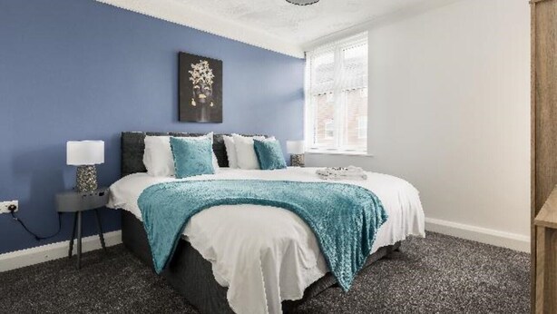 A bedroom with a bed, a bedside table, a lamp, a window, and a wall art piece. - Luxury 3 bedroom apartment (Boscombe)