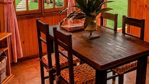A wooden interior with a dining table, chairs, a potted plant, and a view of a garden through a window.