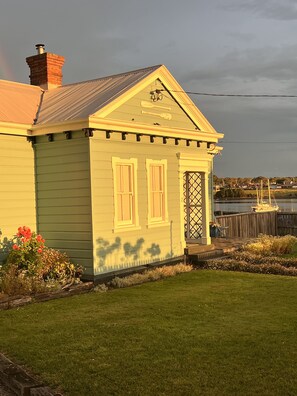A small, single-story house with a gabled roof and a chimney. It features a front door with a lattice design and two windows with shutters. - Lux. Waterfront Cottage, Amazing Water Views /Fire Pit /Central/5km-MTB & Beach (George Town)