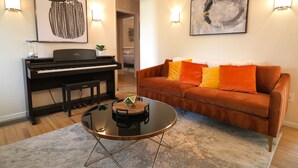 Un saló amb un piano, un sofà marró amb coixins taronja, una taula de café de vidre i dues làmpades penjades a la paret.