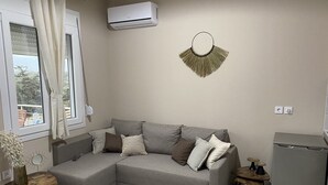 A modern living room with a grey sectional sofa, a small wooden side table, a decorative wall hanging, and a compact refrigerator. - Deatached apartment with shared pool in Tersanas Chania 2 minutes from the beach (Chorafakia)