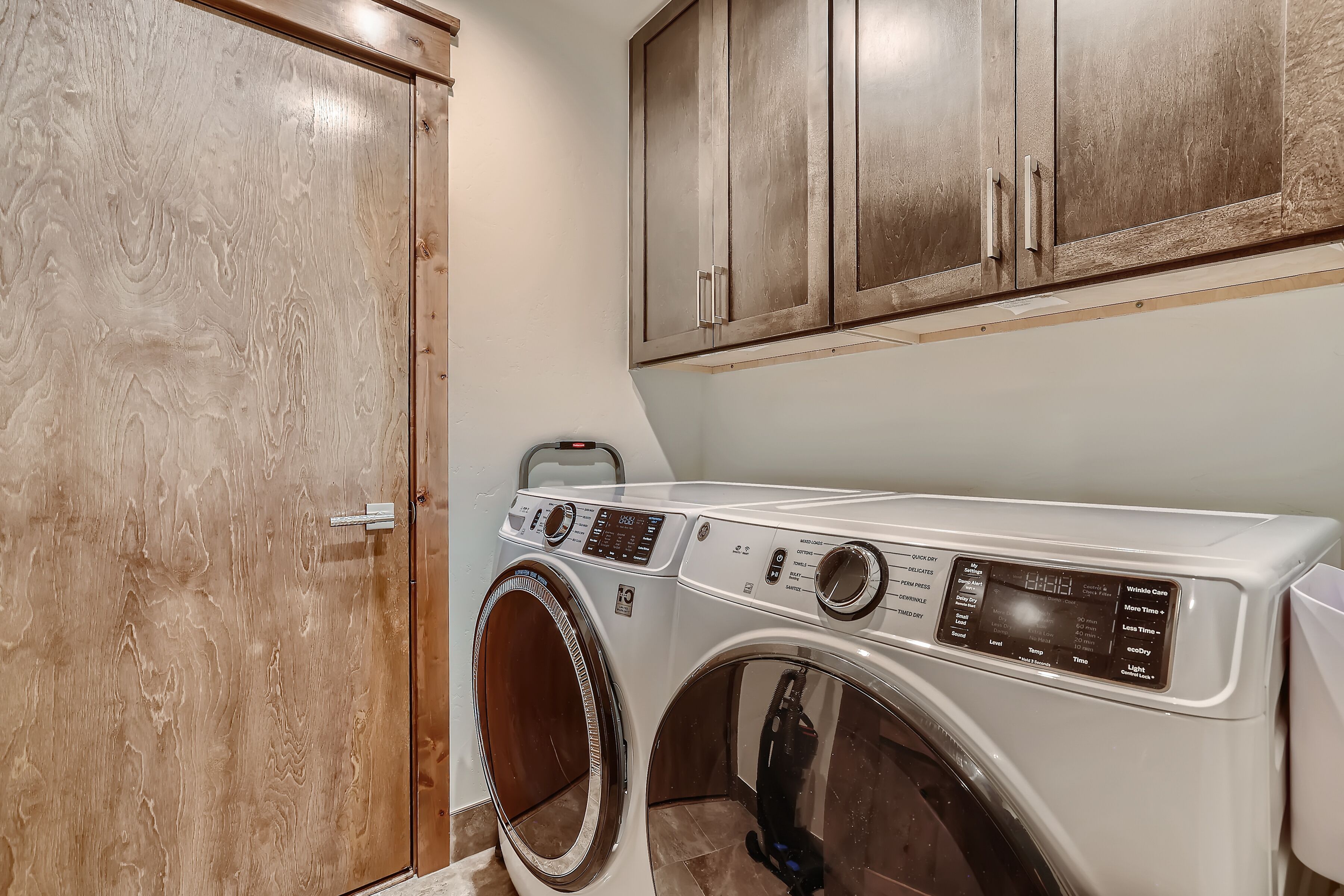 A laundry room with a front-loading washer and dryer, wooden cabinets above, and a door to the left.